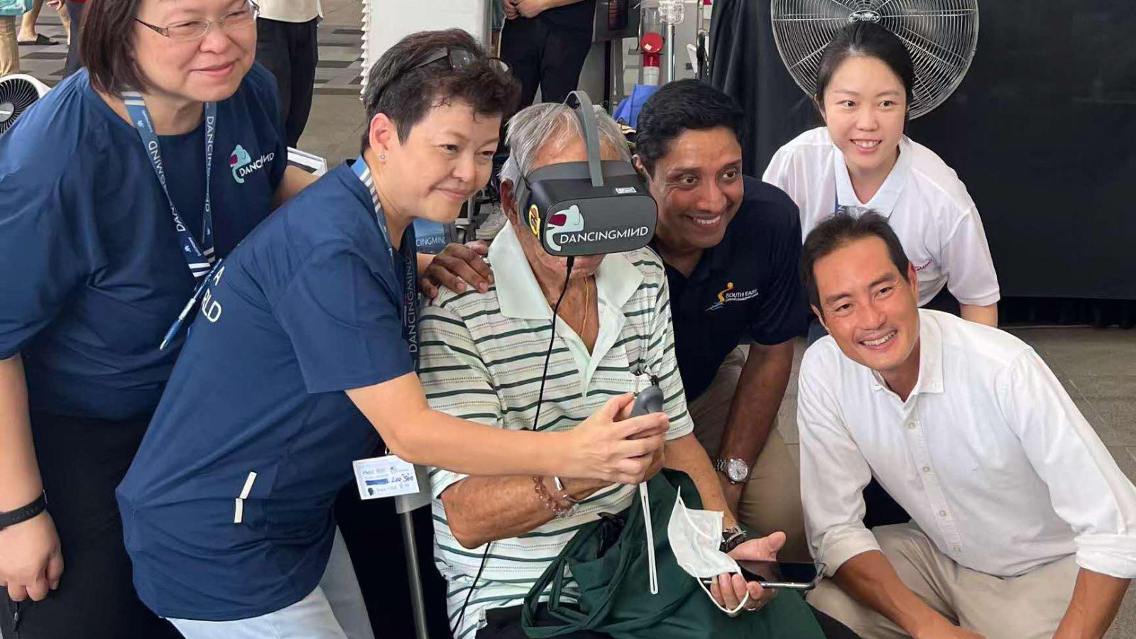 DancingMind founder Jennifer Zhang (top right), Senior Minister of State for Digital Development and Information and Health Tan Kiat How (kneeling, at right), and Minister of State for Culture, Community and Youth and Manpower Dinesh Vasu Dash (third from right) watch as a participant tries the company’s VR rehabilitation technology. 
